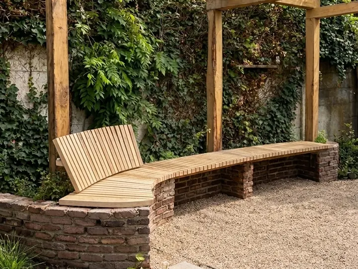 Gebogen houten tuinbank met rugleuning op een gemetseld muurtje, onder een houten pergola tegen een begroeide muur