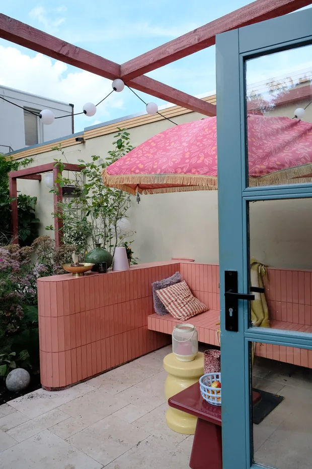Betegelde zithoek op het terras met roze parasol, lichtsnoer en blauwgroene tuindeuren